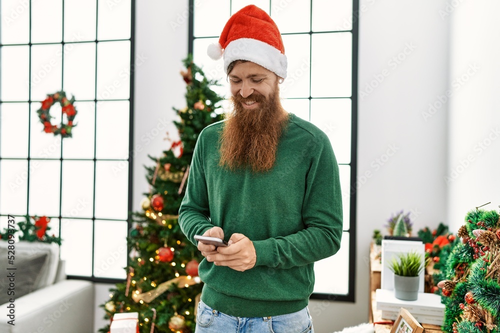 Young redhead man using smartphone standing by christmas tree at home Young redhead man using smartphone standing by christmas tree at home