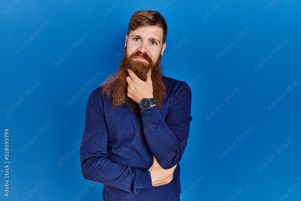 Redhead man with long beard wearing casual blue sweater over blue background looking confident at the camera smiling with crossed arms and hand raised on chin. thinking positive. Redhead man with long beard wearing casual blue sweater over blue background looking confident at the camera smiling with crossed arms and hand raised on chin. thinking positive.