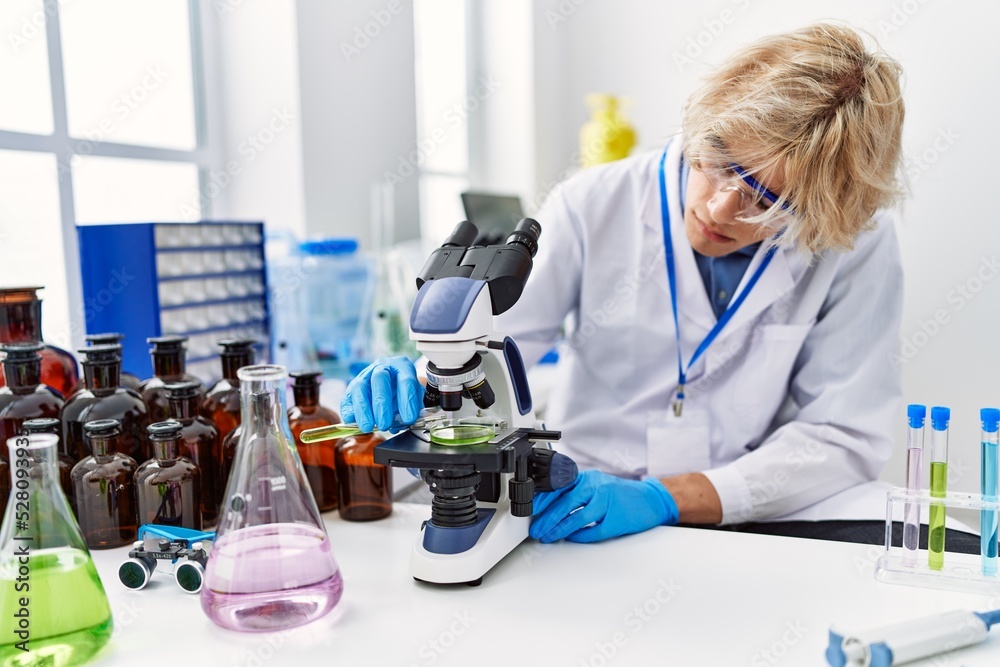 Fototapeta premium Young blond man scientist using microscope pouring liquid on sample at laboratory