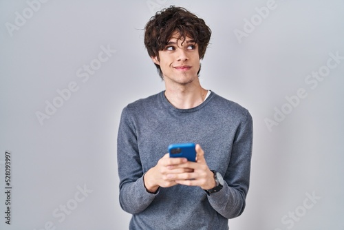 Young man using smartphone typing a message smiling looking to the side and staring away thinking.