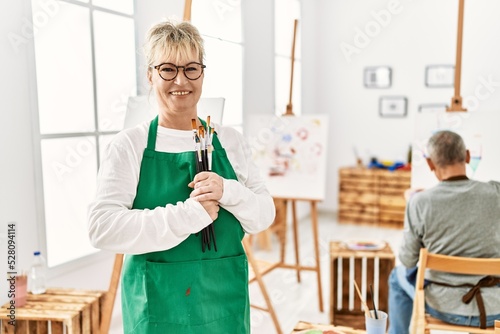 Two senior paint student smiling happy painting at art studio. Woman standing with smile on face holding paintbrushes.