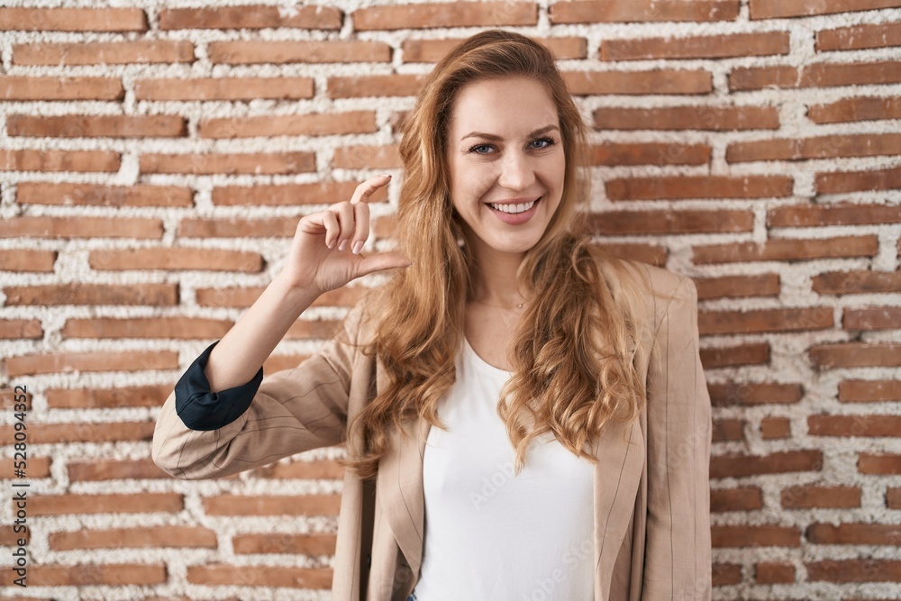 Beautiful blonde woman standing over bricks wall smiling and confident gesturing with hand doing small size sign with fingers looking and the camera. measure concept. Beautiful blonde woman standing over bricks wall smiling and confident gesturing with hand doing small size sign with fingers looking and the camera. measure concept.