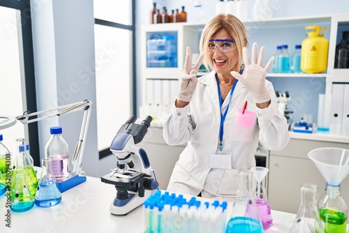 Middle age blonde woman working at scientist laboratory showing and pointing up with fingers number eight while smiling confident and happy.