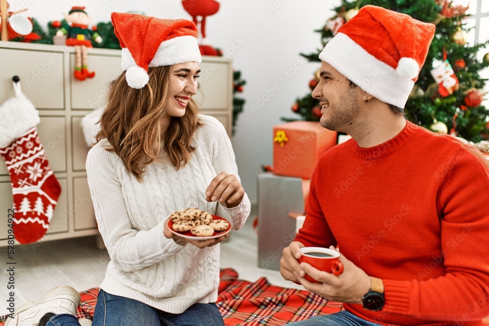 Fototapeta premium Young hispanic couple wearing christmas hat drinking coffee and eating cookies at home.