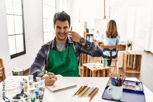 Young artist man at art studio pointing with hand finger to face and nose, smiling cheerful. beauty concept