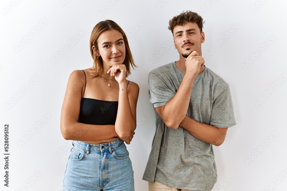 Naklejka premium Young beautiful couple standing together over isolated background looking confident at the camera smiling with crossed arms and hand raised on chin. thinking positive.