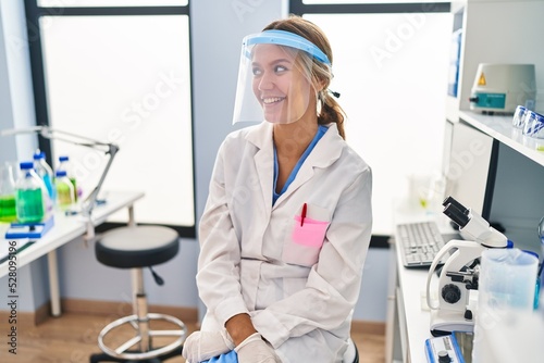 Young blonde woman working at scientist laboratory wearing face mask looking to side  relax profile pose with natural face and confident smile.