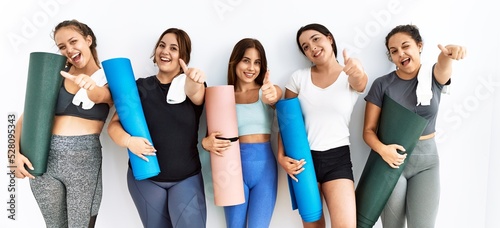 Group of women holding yoga mat standing over isolated background approving doing positive gesture with hand, thumbs up smiling and happy for success. winner gesture.