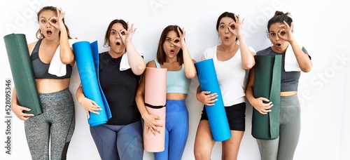 Group of women holding yoga mat standing over isolated background doing ok gesture shocked with surprised face, eye looking through fingers. unbelieving expression.