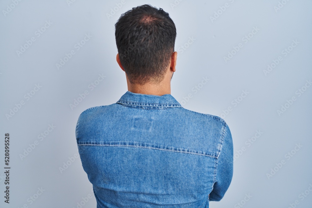 Young hispanic man standing over blue background standing backwards looking away with crossed arms Young hispanic man standing over blue background standing backwards looking away with crossed arms