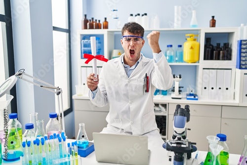 Young hispanic man working at scientist laboratory holding diploma annoyed and frustrated shouting with anger, yelling crazy with anger and hand raised