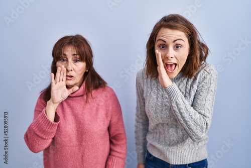 Mother and daughter standing over blue background hand on mouth telling secret rumor, whispering malicious talk conversation