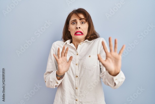 Young beautiful woman standing casual over blue background afraid and terrified with fear expression stop gesture with hands, shouting in shock. panic concept.