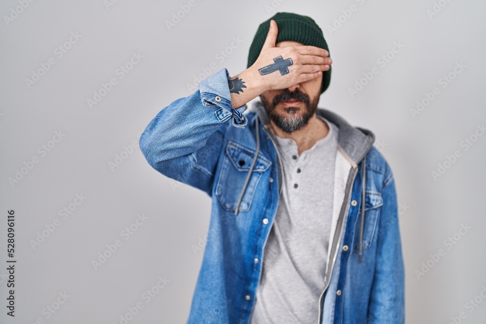 Young hispanic man with tattoos wearing wool cap smiling and laughing with hand on face covering eyes for surprise. blind concept. Young hispanic man with tattoos wearing wool cap smiling and laughing with hand on face covering eyes for surprise. blind concept.