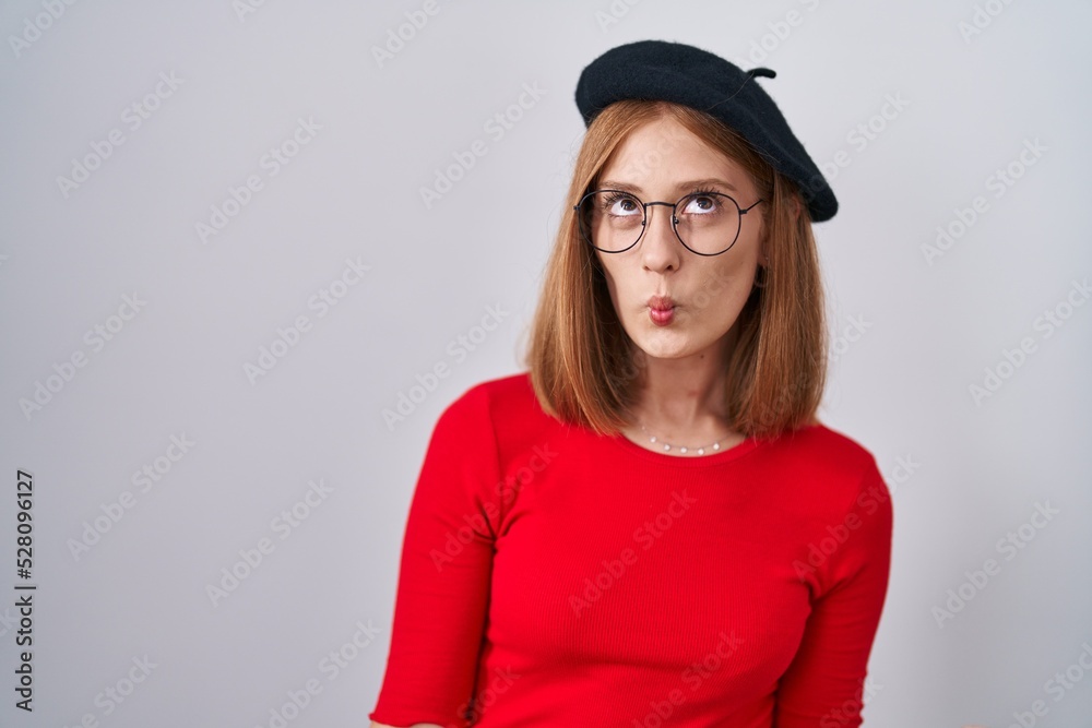 Young redhead woman standing wearing glasses and beret making fish face with lips, crazy and comical gesture. funny expression.