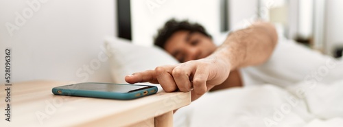 Fotografia Young hispanic man turning off smartphone alarm lying on the bed at bedroom
