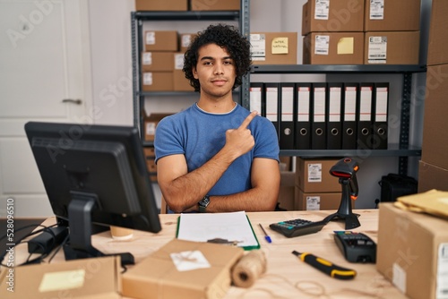 Hispanic man with curly hair working at small business ecommerce pointing with hand finger to the side showing advertisement  serious and calm face