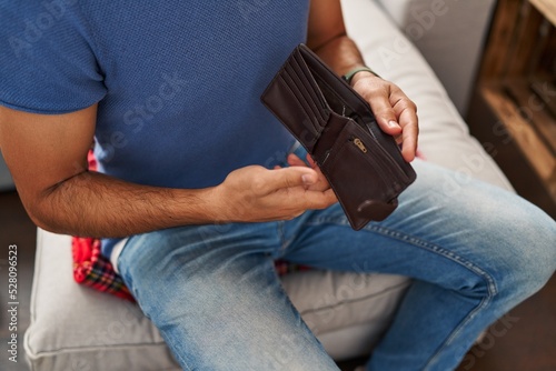 Young hispanic man showing empty wallet sitting on sofa at home