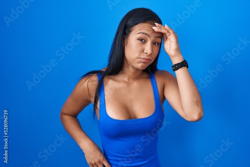 Hispanic woman standing over blue background worried and stressed about a problem with hand on forehead, nervous and anxious for crisis