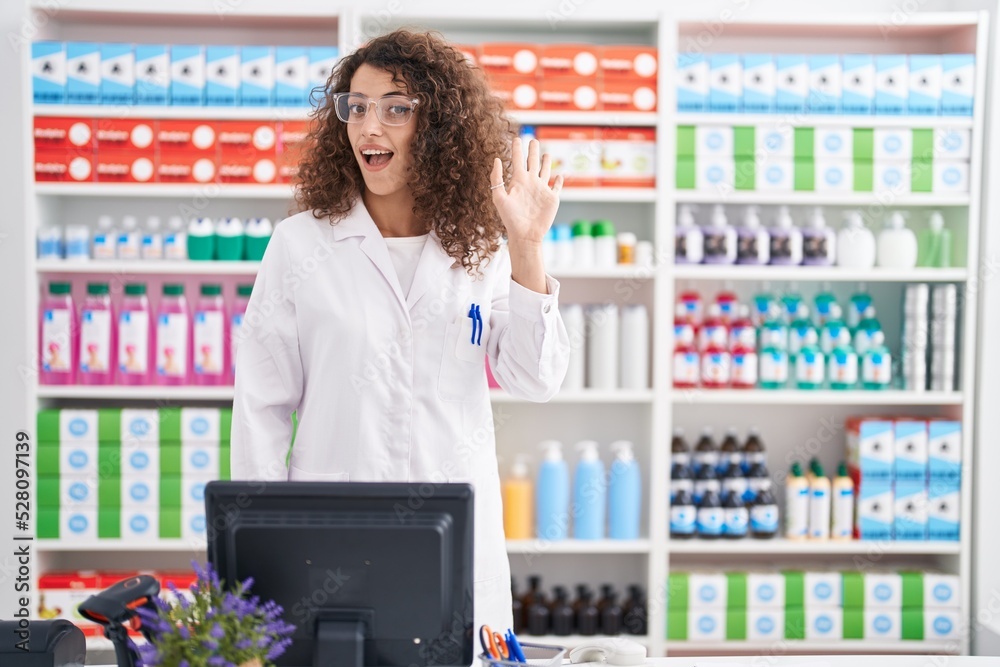 Hispanic woman with curly hair working at pharmacy drugstore waiving saying hello happy and smiling, friendly welcome gesture