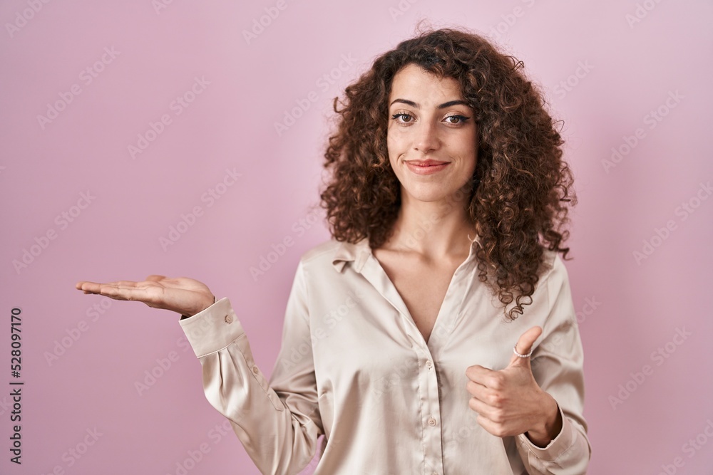 Hispanic woman with curly hair standing over pink background showing palm hand and doing ok gesture with thumbs up, smiling happy and cheerful