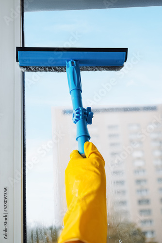 Man in yellow gloves cleaning window with squeegee and spray detergent at home terrace. House cleaning and house chores, domestic hygiene. Window cleaning background with blue sky.