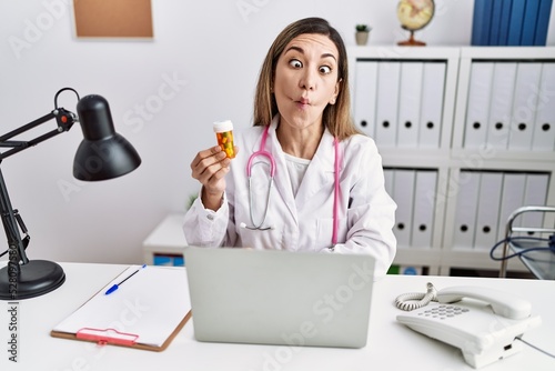 Young hispanic woman wearing doctor uniform holding pills at the clinic making fish face with lips, crazy and comical gesture. funny expression.