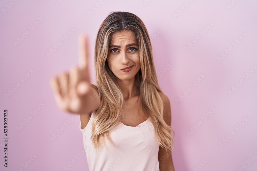Young blonde woman standing over pink background pointing with finger up and angry expression, showing no gesture