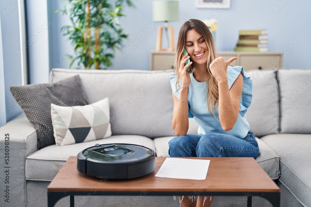 Young hispanic woman sitting at home by vacuum robot speaking on the phone pointing thumb up to the side smiling happy with open mouth Young hispanic woman sitting at home by vacuum robot speaking on the phone pointing thumb up to the side smiling happy with open mouth