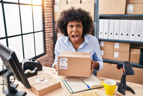 Fotobehang Black woman with curly hair working at small business ecommerce holding box angry and mad screaming frustrated and furious, shouting with anger looking up