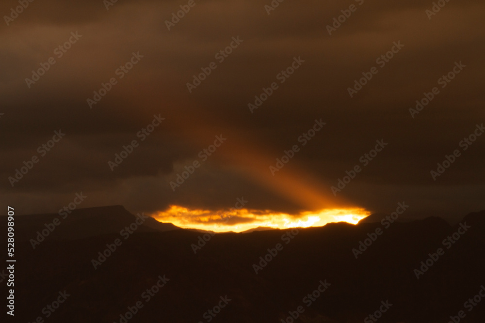 Dark cloudscape. View of the dramatic sunset with dark clouds. The last bright ray of sunlight strikes across the sky and mountains silhouette.
