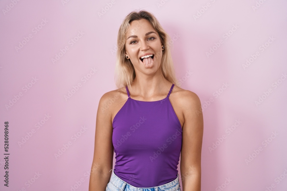 Young blonde woman standing over pink background sticking tongue out happy with funny expression. emotion concept.
