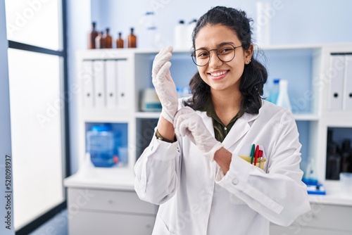 Young latin woman scientist smiling confident wearing gloves at laboratory