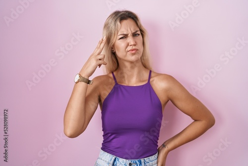 Young blonde woman standing over pink background shooting and killing oneself pointing hand and fingers to head like gun, suicide gesture.