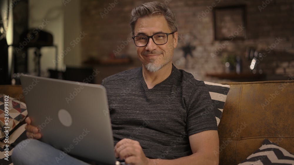 Older man working online with laptop computer at home sitting on couch in living room. Home office, browsing internet. Portrait of happy, mature age, middle age, mid adult man in 50s, smiling. Older man working online with laptop computer at home sitting on couch in living room. Home office, browsing internet. Portrait of happy, mature age, middle age, mid adult man in 50s, smiling.