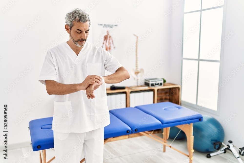 Middle age hispanic therapist man working at pain recovery clinic checking the time on wrist watch, relaxed and confident Middle age hispanic therapist man working at pain recovery clinic checking the time on wrist watch, relaxed and confident