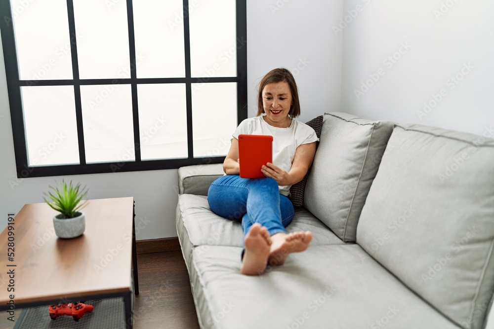 Middle age hispanic woman sitting on the sofa using tablet at home