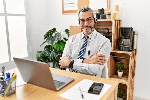 Middle age hispanic business man working at the office wearing operator headset smiling looking to the side and staring away thinking.