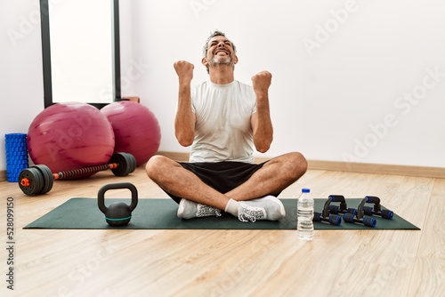 Middle age hispanic man sitting on training mat at the gym very happy and excited doing winner gesture with arms raised, smiling and screaming for success. celebration concept.