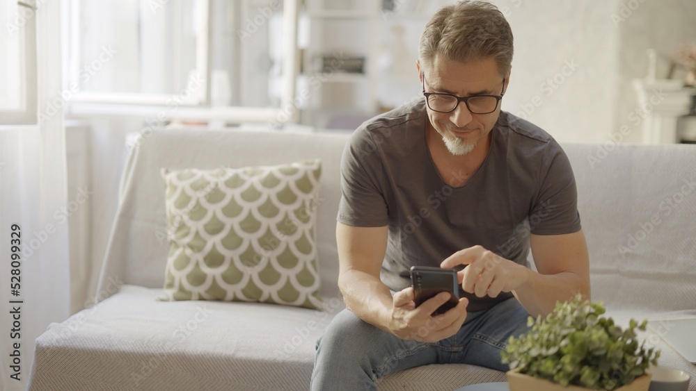 Older man at home, sitting on couch in living room, using phone, checking social media. Portrait of happy, mature age, middle age, mid adult caucasian man in 50s, smiling. Older man at home, sitting on couch in living room, using phone, checking social media. Portrait of happy, mature age, middle age, mid adult caucasian man in 50s, smiling.
