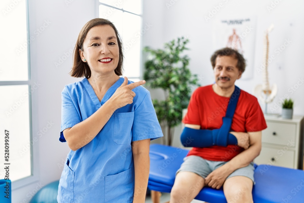 Middle age doctor woman with patient with arm injury at rehabilitation clinic smiling cheerful pointing with hand and finger up to the side Middle age doctor woman with patient with arm injury at rehabilitation clinic smiling cheerful pointing with hand and finger up to the side