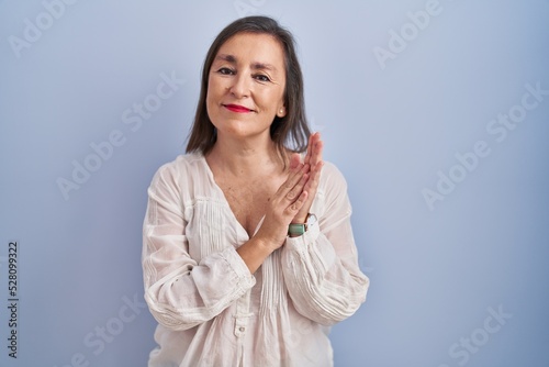Middle age hispanic woman standing over blue background clapping and applauding happy and joyful, smiling proud hands together © Krakenimages.com
