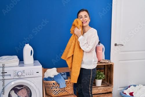 Middle age woman washing clothes touching softener sweater with face at laundry room