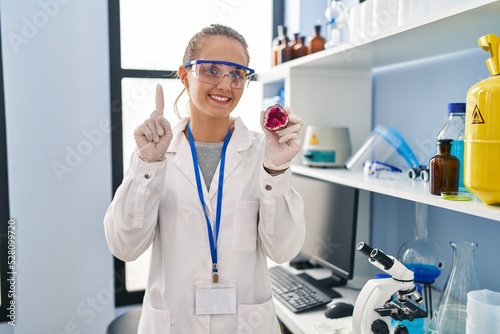 Young woman working at scientist laboratory holding geode smiling with an idea or question pointing finger with happy face  number one