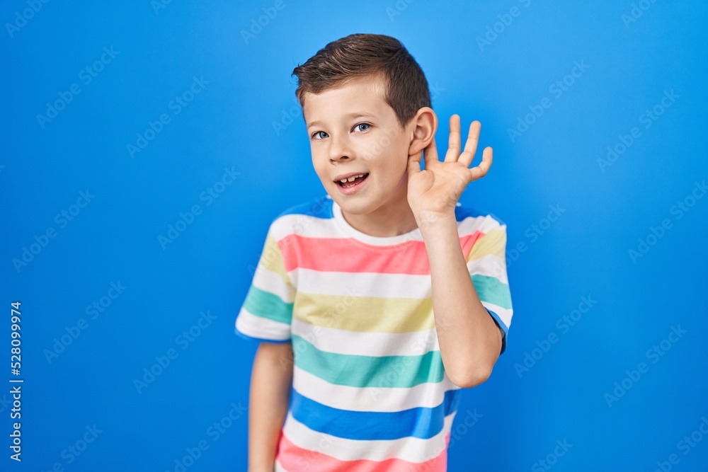 Young caucasian kid standing over blue background smiling with hand over ear listening an hearing to rumor or gossip. deafness concept. Young caucasian kid standing over blue background smiling with hand over ear listening an hearing to rumor or gossip. deafness concept.