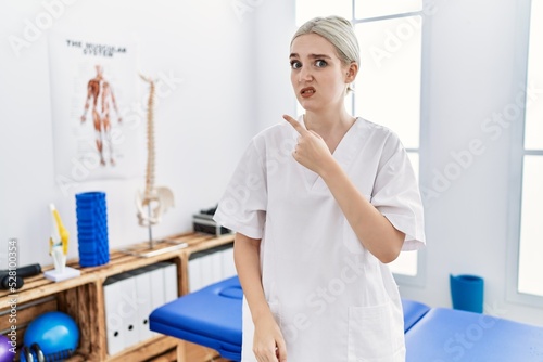 Young caucasian woman working at pain recovery clinic pointing aside worried and nervous with forefinger  concerned and surprised expression