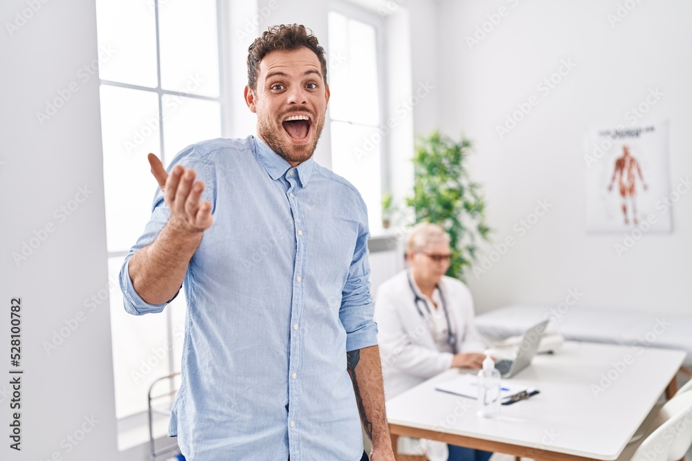 Hispanic man at doctor clinic celebrating achievement with happy smile and winner expression with raised hand