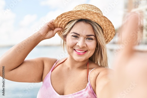 Young hispanic woman wearing summer hat make selfie by the smartphone at seaside