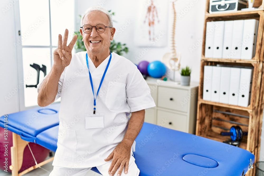 Senior physiotherapy man working at pain recovery clinic showing and pointing up with fingers number three while smiling confident and happy. Senior physiotherapy man working at pain recovery clinic showing and pointing up with fingers number three while smiling confident and happy.