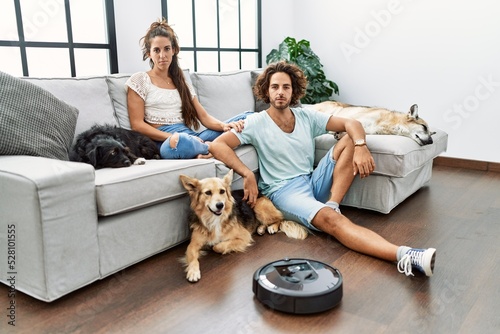Young hispanic couple relaxing at home by vacuum robot thinking attitude and sober expression looking self confident